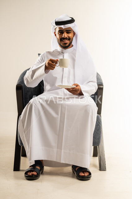النظر الى الكاميرا بإيماءات السرور والسعادة, قضاء وقت الفراغ باحتساء المشروب المفضل, الجلوس والاستمتاع بالمشروبات الساخنة والأعشاب الشتوية الدافئة, بورتريه لشاب عربي خليجي كويتي يرتدي الثوب التقليدي والغتره يجلس على كرسي مريح يقوم باحتساء القهوة, خلفية بيضاء