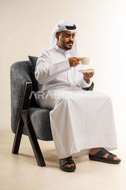 الجلوس والاستمتاع بالمشروبات الساخنة والأعشاب الشتوية الدافئة, قضاء وقت الفراغ باحتساء المشروب المفضل, بورتريه لشاب عربي خليجي كويتي يرتدي الثوب التقليدي والغتره يجلس على كرسي مريح يقوم بشرب القهوة, خلفية بيضاء