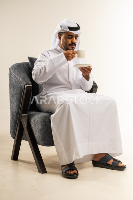 قضاء وقت الفراغ باحتساء المشروب المفضل, الجلوس والاستمتاع بالمشروبات الساخنة والأعشاب الشتوية الدافئة, بورتريه لشاب عربي خليجي كويتي يرتدي الثوب التقليدي والغتره يجلس على كرسي مريح يقوم باحتساء القهوة, خلفية بيضاء