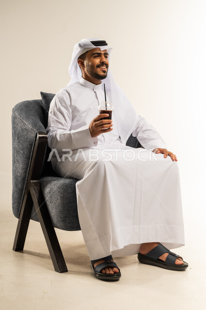 Trying to relieve the summer heat with a refreshing drink, enjoying delicious healthy drinks, portrait of a young Gulf Arab Kuwaiti man wearing a traditional thobe sitting on a comfortable chair enjoying drinking cold natural juice, gestures of pleasure and happiness, white background
