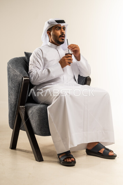 إيماءات الذهول والصدمة, محاولة التخفيف من حرارة الصيف بالمشروب المنعش، الاستمتاع بالمشروبات المفيدة اللذيذة، بورتريه لشاب عربي خليجي كويتي يرتدي الثوب التقليدي والغتره يجلس على كرسي مريح ويستمتع بشرب العصير الطبيعي البارد, خلفية بيضاء