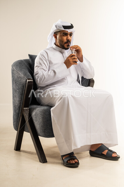 Trying to relieve the summer heat with a refreshing drink, enjoying delicious healthy drinks, portrait of a young Gulf Arab Kuwaiti man wearing a traditional thobe sitting on a comfortable chair enjoying drinking cold natural juice, gestures of pleasure and happiness, white background