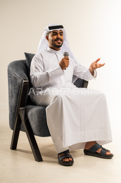 ممارسة موهبة الغناء، تسجيل مقطع صوتي غنائي، بورتريه لشاب عربي خليجي كويتي يرتدي الثوب التقليدي والغتره يجلس على كرسي مريح يمسك بيده المايكروفون يغني بإيماءات الحماس والسرور، تقديم العروض الصوتية، تعابير الاندماج والتفاعل مع الموسيقا، خلفية بيضاء