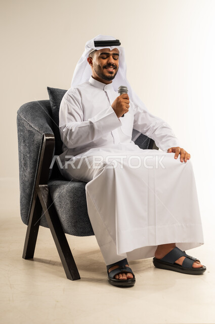 تسجيل مقطع صوتي غنائي، ممارسة موهبة الغناء، بورتريه لشاب عربي خليجي كويتي يرتدي الثوب التقليدي والغتره يجلس على كرسي مريح يمسك بيده المايكروفون يغني بإيماءات الحماس والسرور، تقديم العروض الصوتية، تعابير الاندماج والتفاعل مع الموسيقا، خلفية بيضاء