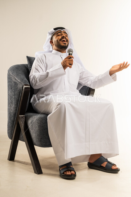 تعابير الاندماج والتفاعل مع الموسيقا، تسجيل مقطع صوتي غنائي، بورتريه لشاب عربي خليجي كويتي يرتدي الثوب التقليدي والغتره يجلس على كرسي مريح يمسك بيده المايكروفون يغني بإيماءات الحماس والسرور، تقديم العروض الصوتية، ممارسة موهبة الغناء، خلفية بيضاء