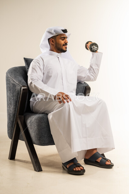 Exercising and lifting weights with dumbbells, portrait of a young Arab Gulf Kuwaiti man wearing traditional dress sitting on a comfortable chair holding heavy iron weights in his hand, maintaining health, physical fitness and physical agility, gestures of pleasure and joy, white background