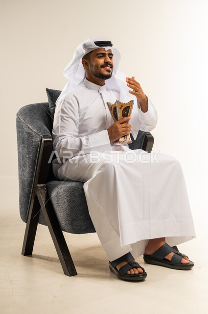 ايماءات الفرح والسرور, العادات و التقاليد العريقه في الكويت, استخدام خشب العود في الاعياد والمناسبات، بورتريه لشاب عربي خليجي كويتي يرتدي الثوب التقليدي والغتره يجلس على كرسي مريح يقوم بالتطيب و التعطر من البخور( المبخرة ), خلفية بيضاء