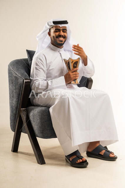 Kuwait's ancient customs and traditions, looking at the camera with gestures of joy and happiness, using oud wood on holidays and occasions, portrait of a young Gulf Arab Kuwaiti man wearing traditional dress sitting on a comfortable chair applying perfume and incense (incense burner), white background
