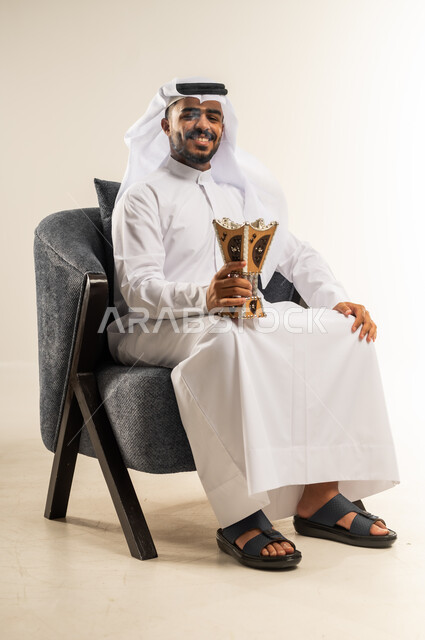 استخدام خشب العود في الاعياد والمناسبات، بورتريه لشاب عربي خليجي كويتي يرتدي الثوب التقليدي والغتره يجلس على كرسي مريح يقوم بالتطيب و التعطر من البخور( المبخرة ), العادات و التقاليد العريقه في الكويت, النظر إلى الكاميرا بايماءات الفرح والسرور, خلفية بيضاء