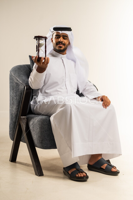 مفهوم إدارة الأعمال وتنظيم الجدول الزمني, القيام بمراقبة توقيت الرمل, إدراك قيمة اللحظات الثمينة, بورتريه لشاب عربي خليجي كويتي يرتدي الثوب التقليدي والغتره يجلس على كرسي مريح يمسك بيده الساعة الرملية الزجاجية (البنكام) ينظر اليها بإيماءات السرور والفرح, خلفية بيضاء
