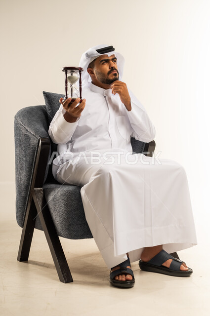 إدراك قيمة اللحظات الثمينة, القيام بمراقبة توقيت الرمل, مفهوم إدارة الأعمال وتنظيم الجدول الزمني, بورتريه لشاب عربي خليجي كويتي يرتدي الثوب التقليدي والغتره يجلس على كرسي مريح يمسك بيده الساعة الرملية الزجاجية (البنكام) يضع يده على ذقنه بتعابير التفكير, خلفية بيضاء
