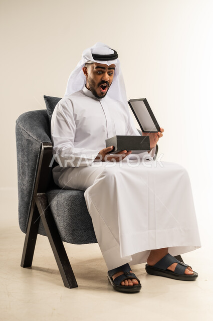 تقديم الهدايا بالاعياد والمناسبات الخاصة، مفهوم الشكر والامتنان، التعبير عن الحب والتقدير والاحترام، بورتريه لشاب عربي خليجي عماني يرتدي الثوب التقليدي والغتره يجلس على كرسي مريح يحمل بيديه صندوق هدية, ايماءات الفرح والسرور، خلفية بيضاء