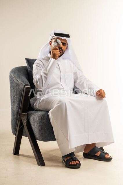 مفهوم الفحص والاستكشاف، توضيح الصورة والرؤية عن قرب، بورتريه لشاب عربي خليجي كويتي يرتدي الثوب التقليدي والغتره يجلس على كرسي مريح يحمل بيده عدسة مكبرة يضعها على عينه بإيماءات التركيز والتمعن، خلفية بيضاء