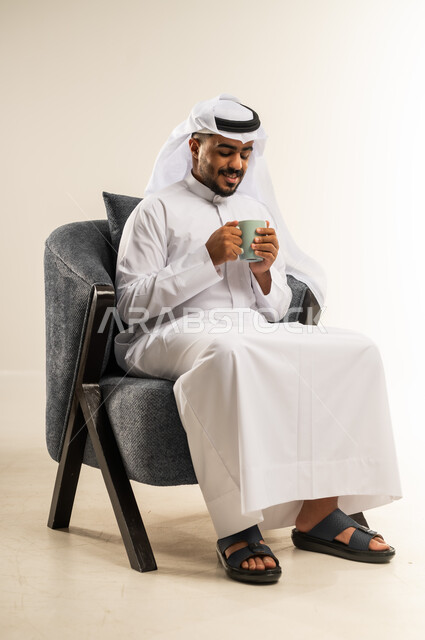 Sitting and enjoying hot drinks and warm winter herbs, spending leisure time sipping your favorite drink, portrait of a young Arab Gulf Omani man wearing traditional dress sitting on a comfortable chair drinking coffee with gestures of happiness and joy, white background