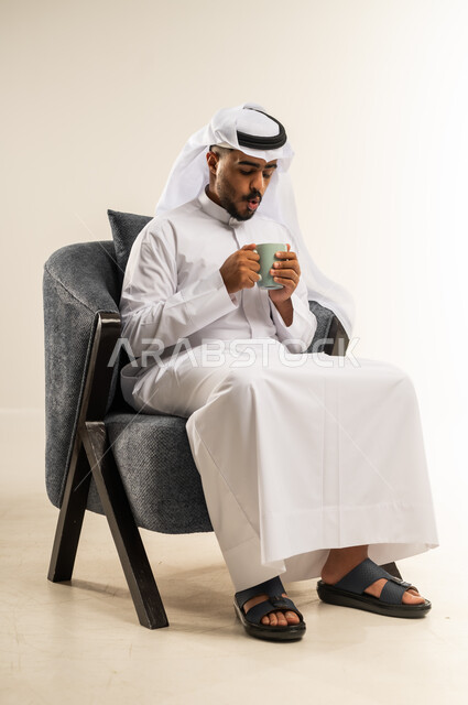قضاء وقت الفراغ باحتساء المشروب المفضل, الجلوس والاستمتاع بالمشروبات الساخنة والأعشاب الشتوية الدافئة, بورتريه لشاب عربي خليجي كويتي يرتدي الثوب التقليدي والغتره يجلس على كرسي مريح يقوم بشرب القهوة, خلفية بيضاء