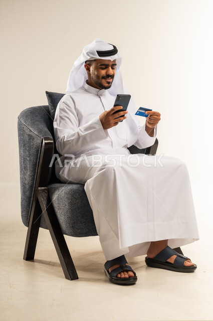 سهولة الشراء والدفع عن طريق بطاقة الإئتمان البنكية، استخدام الأجهزة والتقنيات الحديثة, بورتريه لشاب عربي خليجي كويتي يجلس على كرسي مريح يرتدي الثوب التقليدي والغتره يمسك بيده بطاقة الصراف و الهاتف المحمول, خلفية بيضاء