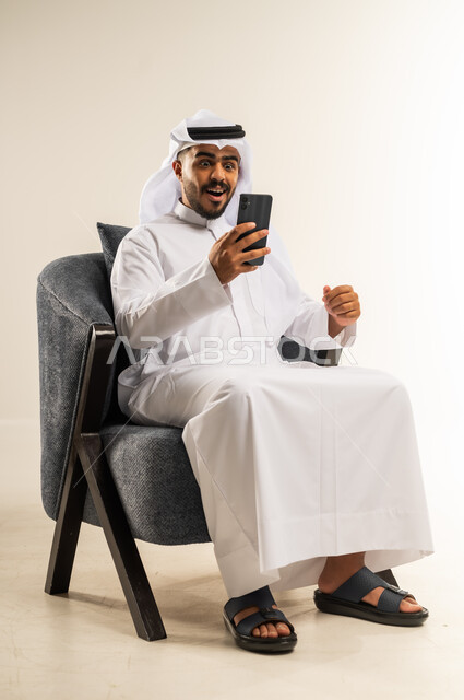 التواصل والدردشة مع الأهل والأصدقاء عبر تطبيقات الجوال, تصفح مواقع التواصل الاجتماعي, بورتريه لشاب عربي خليجي كويتي يرتدي الثوب التقليدي والغتره يجلس على كرسي مريح يرفع قبضة يده بتعابير الفوز و الربح, استخدام التقنيات الحديثة المتطورة, خلفية بيضاء
