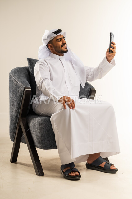 استخدام التقنيات الحديثة المتطورة, اجراء مكالمة فيديو مع الأهل والأصدقاء عبر الجوال, تصفح مواقع التواصل الاجتماعي, بورتريه لشاب عربي خليجي كويتي يرتدي الثوب التقليدي والغتره يجلس على كرسي مريح يمسك الهاتف المحمول, خلفية بيضاء