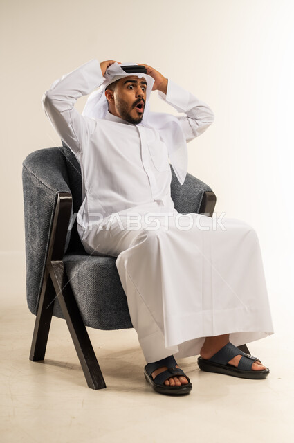التعجب والإستغراب والصدمة، بورتريه لرجل عربي خليجي كويتي يرتدي الثوب التقليدي والغتره يجلس على كرسي مريح يضع يده على رأسه بإيماءات الدهشة والذهول، تلقي اخبار سيئة غير متوقعة، خلفية بيضاء