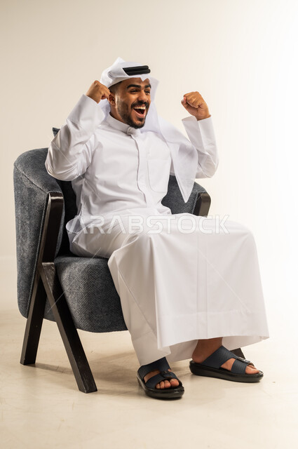 الانتصار والفوز والنجاح، مفهوم الانجاز وتحقيق الاهداف، بورتريه لشاب عربي خليجي كويتي يرتدي الثوب التقليدي والغتره يجلس على كرسي مريح يرفع قبضة يديه للاعلى بإيماءات الفرح والسرور, الشعور بالسعادة والقوة, خلفية بيضاء