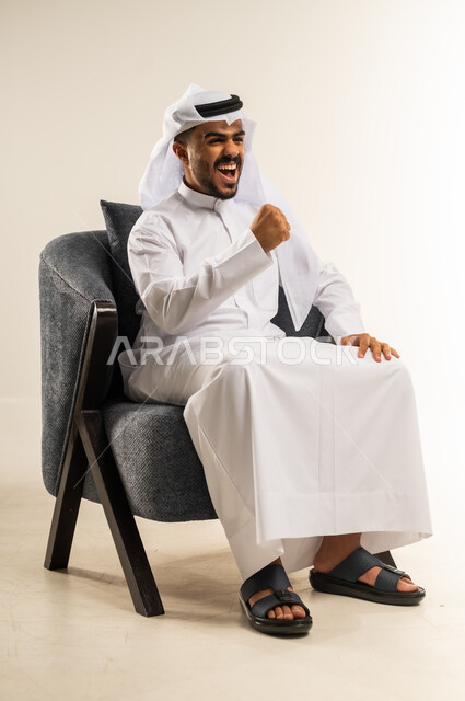 مفهوم الانجاز وتحقيق الاهداف، الانتصار والفوز والنجاح، بورتريه لشاب عربي خليجي كويتي يرتدي الثوب التقليدي والغتره يجلس على كرسي مريح يرفع قبضة يده للاعلى بإيماءات الفرح والسرور, الشعور بالسعادة والقوة, خلفية بيضاء