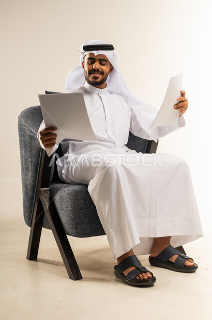 Proofreading notes, preparing work reports, portrait of a smiling young Kuwaiti Gulf Arab man wearing traditional thobe sitting on a comfortable chair holding a bunch of white papers, reviewing and following up on office plans and projects with focus gestures, white background