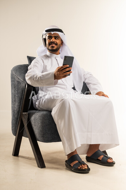الإصغاء والاستمتاع بإستخدام الهيدفون، تصفح مواقع التواصل الاجتماعي, اجراء مكالمة فيديو مع الأهل والأصدقاء عبر الجوال, بورتريه لشاب عربي خليجي كويتي يرتدي ثوب التقليدي والغتره يجلس على كرسي مريح يمسك الهاتف المحمول, استخدام التقنيات الحديثة المتطورة, خلفية بيضاء