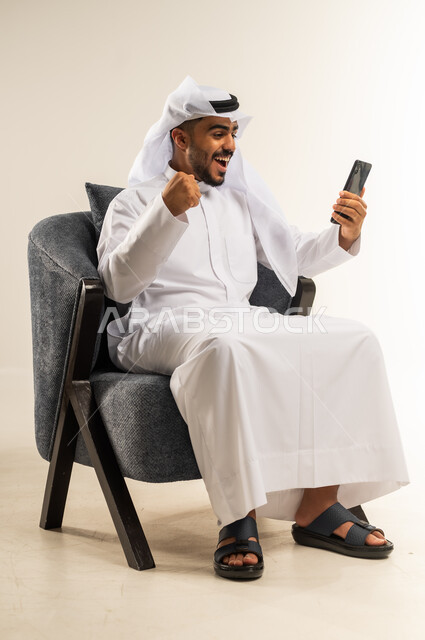 Browsing social networking sites, communicating and chatting with family and friends via mobile applications, portrait of a young Kuwaiti Gulf Arab man wearing traditional dress sitting on a comfortable chair raising his fist with expressions of victory and profit, using modern advanced technologies, background White
