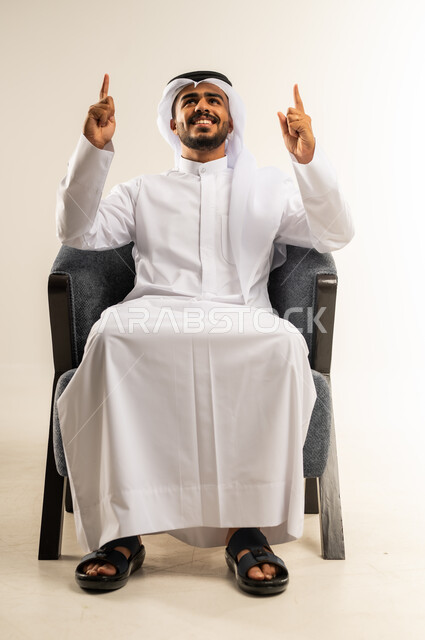 Gestures indicating this place (here), specifying a specific location, confirming that something is happening at this moment, portrait of a Kuwaiti Gulf Arab man wearing traditional thobe and ghutra pointing his index fingers upwards, gestures of pleasure and happiness, white background
