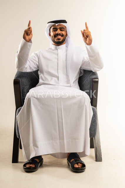 تأكيد حدوث شيء هذه اللحظة, حركات تدل على هذا المكان (هنا)، تحديد موقع معين، بورتريه لرجل عربي خليجي كويتي يرتدي الثوب التقليدي والغترة يشير بإصابع السبابة نحو الأعلى, النظر إلى الكاميرا بايماءات السرور، خلفية بيضاء