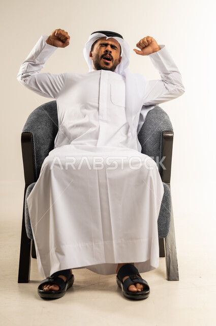 الشعور بالتعب والحاجة إلى النوم, الإحساس بالكسل والخمول و الإرهاق، بورتريه لرجل عربي خليجي كويتي يرتدي الثوب التقليدي والغتره يجلس على كرسي مريح يرفع يديه للاعلى بإيماءات التثاؤب والنعاس, خلفية بيضاء