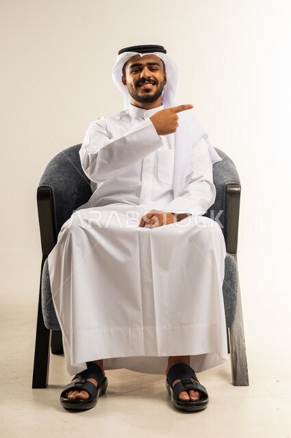 الاشارة باصبع السبابة الى موقع معين، إشارات وحركات يدين تدل على هذا المكان (هنا)، بورتريه لرجل عربي خليجي كويتي يرتدي الثوب التقليدي والغتره ينظر الى الكاميرا بايماءات السعادة والسرور، مفهوم الأناقة والاهتمام بالمظهر الخارجي، خلفيه بيضاء