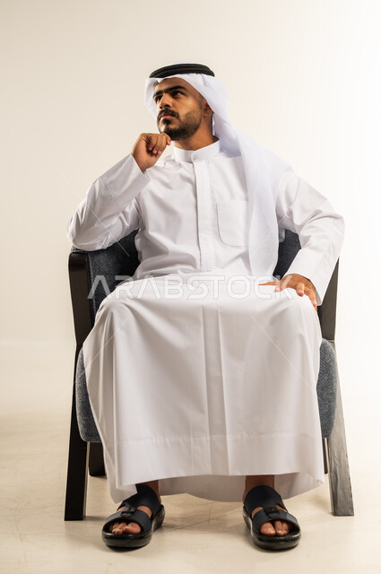التمعن والتأمل في موضوع معين، بورتريه لرجل عربي خليجي كويتي يرتدي الثوب التقليدي والغتره يجلس على كرسي مريح ويضع يده على ذقنه، النظر الى شيء ما إيماءات الملل واللامبالاة، التفكير والحيرة والتركيز في حل المشكلات، خلفية بيضاء