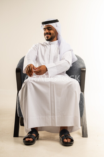 The concept of elegance and masculinity, interest in external appearance, lifting and carrying a certain item, portrait of a young Kuwaiti Gulf Arab man wearing traditional dress sitting on a comfortable chair trying to hold something, gestures of happiness and pleasure, white background