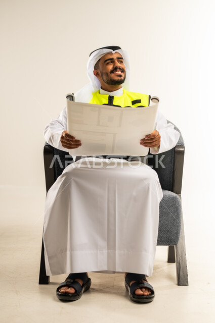 التدقيق والإشراف على مخططات المشروع، إيماءات البهجة والسرور، مهن ووظائف هندسية في الكويت، بورتريه لمهندس عربي خليجي كويتي يرتدي الثوب التقليدي وسترة الحماية الخاصة بالعمل يجلس على كرسي مريح يمسك بيده مخطط هندسي، خلفية بيضاء