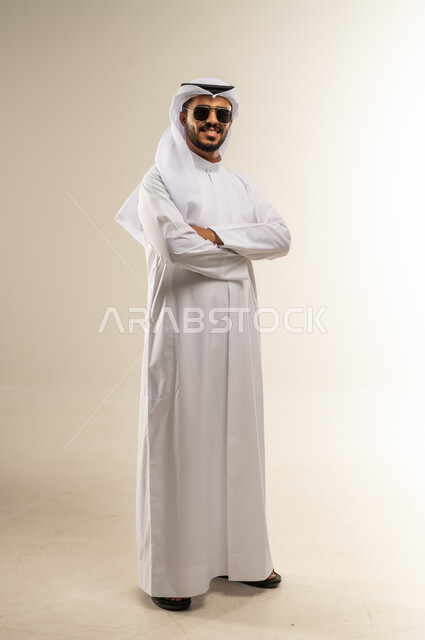 مفهوم الرجولة والثقة بالنفس، الاهتمام بالمظهر الخارجي والأناقة، بورتريه لرجل عربي خليجي كويتي يرتدي الثوب التقليدي  يضع نضارة شمسية و يقف بايدي مكتوفة، الوقوف باستقامة وثبات، النظر الى الكاميرا بايماءات السرور والفرح, صورة الجسم بالطول الكامل، خلفية بيضاء