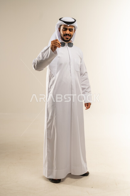 الاهتمام بالمظهر الخارجي والأناقة، مفهوم الرجولة والثقة بالنفس، بورتريه لرجل عربي خليجي كويتي يرتدي الثوب التقليدي يقف بايدي مكتوفة ويمسك بيده نضارة شمسية، الوقوف باستقامة وثبات، ايماءات السرور والفرح, صورة الجسم بالطول الكامل، خلفية بيضاء
