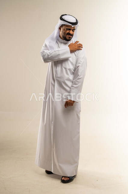 الأوجاع والآلام العصبية والعضلية، طلب المساعده والحاجه للعلاج، التهاب العضلات والمفاصل، بورتريه لرجل عربي خليجي كويتي يرتدي الثوب التقليدي يضع يده على كتفه بايماءات الألم والتعب والأرهاق، صورة الجسم الطول الكامل, خلفية بيضاء