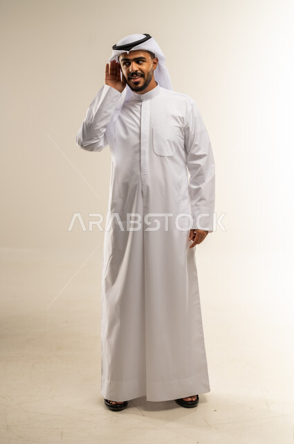 الحاجة لمراجعة طبيب الأذن لتركيب السماعات الطبية, إيماءات التركيز والاستماع لفهم الكلام بشكل واضح، بورتريه لرجل عربي خليجي كويتي يرتدي الثوب التقليدي يمسك أذنه بيده ويعاني من ضعف السمع، الجسم بالطول الكامل، خلفية بيضاء