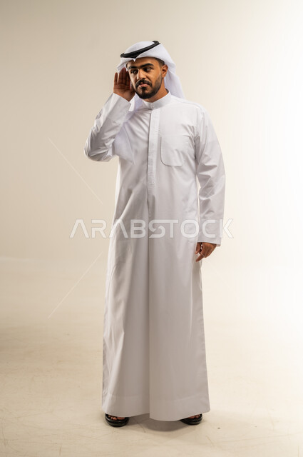 الحاجة لمراجعة طبيب الأذن لتركيب السماعات الطبية, إيماءات التركيز والاستماع لفهم الكلام بشكل واضح، بورتريه لرجل عربي خليجي كويتي يرتدي الثوب التقليدي يمسك أذنه بيده ويعاني من ضعف السمع، الجسم بالطول الكامل، خلفية بيضاء