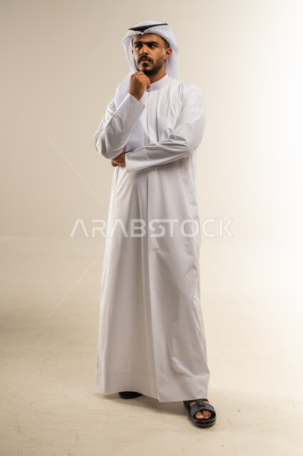 وضع اليد تحت الذقن بتعابير القلق وعدم اتخاذ القرار، إيماءات التفكير العميق والحيرة، بورتريه لرجل عربي خليجي كويتي يرتدي الثوب التقليدي يقف وينظر إلى مكان بعيد ويخطط لشيء ما، صورة الجسم بالطول الكامل، خلفية بيضاء