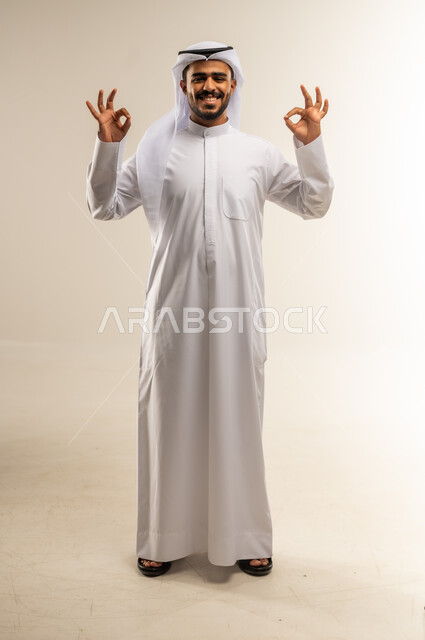 التعبير عن الإعجاب والتقدير، الشعور بالرضا والاستحسان، رفع اليدين بعلامات الجودة والقبول، الامتياز والعلامة الكاملة، بورتريه لرجل عربي خليجي كويتي مبتسم يرتدي الثوب التقليدي والغتره ينظر إلى الكاميرا بإيماءات السعادة والسرور، الجسم بالطول الكامل، خلفية بيضاء