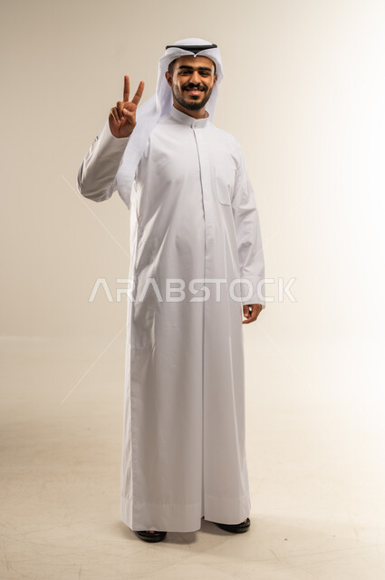 النجاح وتحقيق الأهداف، مفهوم السعادة بالوصول إلى الغايات، بورتريه لرجل عربي خليجي كويتي يرتدي الثوب التقليدي يرفع يده باشارة النصر والقوة، إيماءات السرور بالفوز والانتصار، صورة الجسم بالطول الكامل, خلفية بيضاء