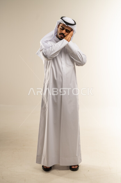 الشعور بالتعب والحاجة إلى الراحة, الإحساس بالكسل والخمول و الإرهاق، بورتريه لرجل عربي خليجي كويتي يرتدي الثوب التقليدي يضع يده تحت راسه بايماءات النوم, صورة الجسم بالطول الكامل, خلفية بيضاء
