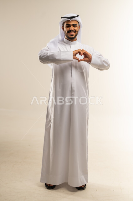 التعبير عن الاعجاب والمحبة، بورتريه لرجل عربي خليجي كويتي يرتدي الثوب التقليدي يرفع يديه باشارة قلب الحب، إيماءات السعادة والسرور، صورة الجسم بالطول الكامل, خلفية بيضاء
