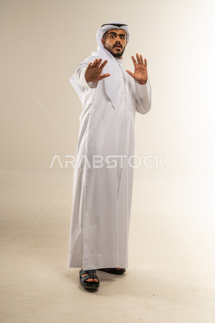 القلق والاحتياط والابتعاد عن شيء ما, حماية النفس من ضرر خارجي, بورتريه لرجل عربي خليجي كويتي يرتدي الثوب التقليدي يرفع يديه للاعلى بإيماءات التوقف وعدم الاقتراب، مفهوم الحذر والصدمة، صورة الجسم بالطول الكامل، خلفية بيضاء