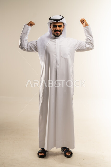 التمتع بالبنية الجسدية القوية، بورتريه لرجل عربي خليجي كويتي يرتدي الثوب التقليدي ويقوم بعرض عضلات يديه، إيماءات وجه ويدين تدل على القوة العضلية، اللياقة البدنية والجسم الصحي المتين، صورة الجسم الطول الكامل، خلفية بيضاء