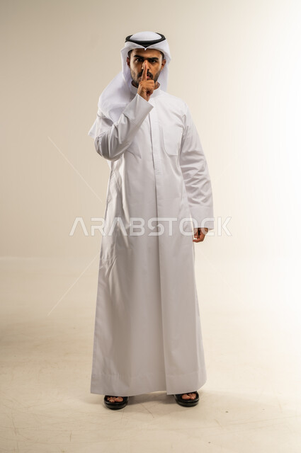التوقف عن الكلام و الأمر بالصمت والسكوت، بورتريه لشاب عربي خليجي كويتي يرتدي الثوب التقليدي ينظر الى الكاميرا يرفع اصبعه الى فمه بإيماءات طلب الهدوء وعدم الازعاج, صورة الجسم بالطول الكامل، خلفية بيضاء