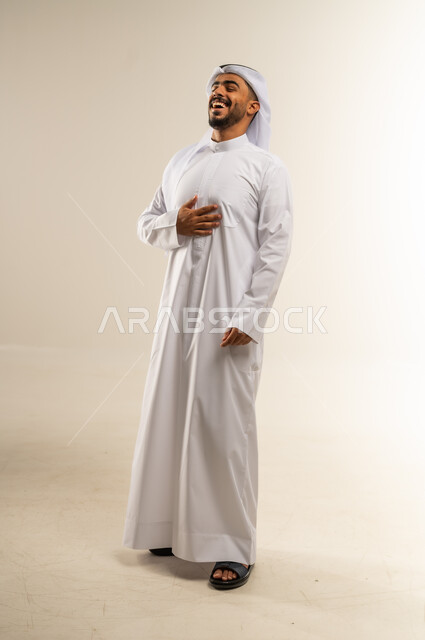 الفرح والسعادة العارمة وقضاء أوقات ممتعة، إغماض العينين والضحك بشدة، سماع خبر ما مضحك، المزاح وإلقاء النكات، بورتريه لرجل عربي خليجي كويتي مبتسم يرتدي الثوب التقليدي والغتره يمسك بطنه ويضحك، الجسم بالطول الكامل، خلفية بيضاء