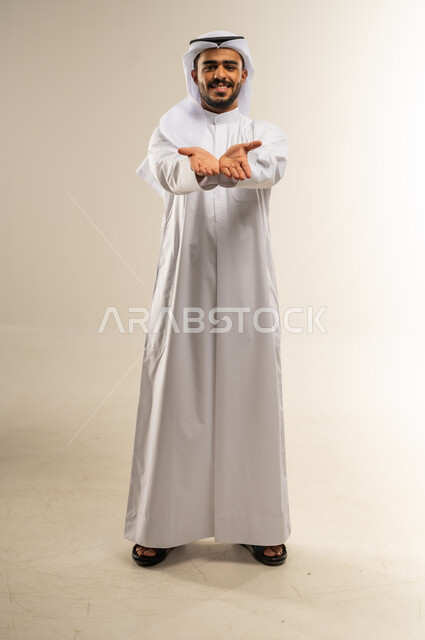 فتح الكفين ممدودة للأمام، تقديم الضيافة أو إعطاء شيء ما، الأناقة والوسامة والرجولة، بورتريه لرجل عربي خليجي كويتي مبتسم يرتدي الثوب التقليدي والغتره ينظر إلى الكاميرا بإيماءات السعادة والسرور، الجسم بالطول الكامل، خلفية بيضاء